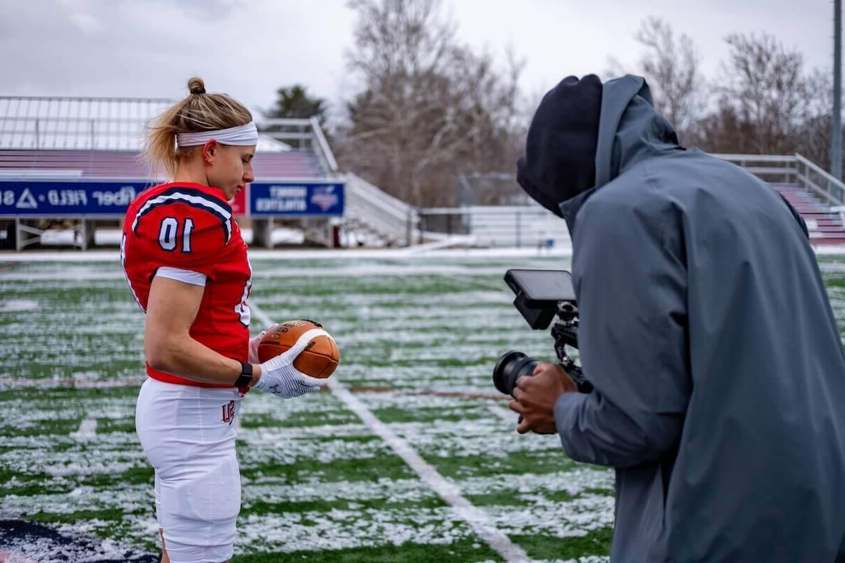 哈利·范·沃里斯身穿全球最大的博彩平台的红色橄榄球制服，在橄榄球场上拿着一个足球，而一名摄像师正在录制视频.